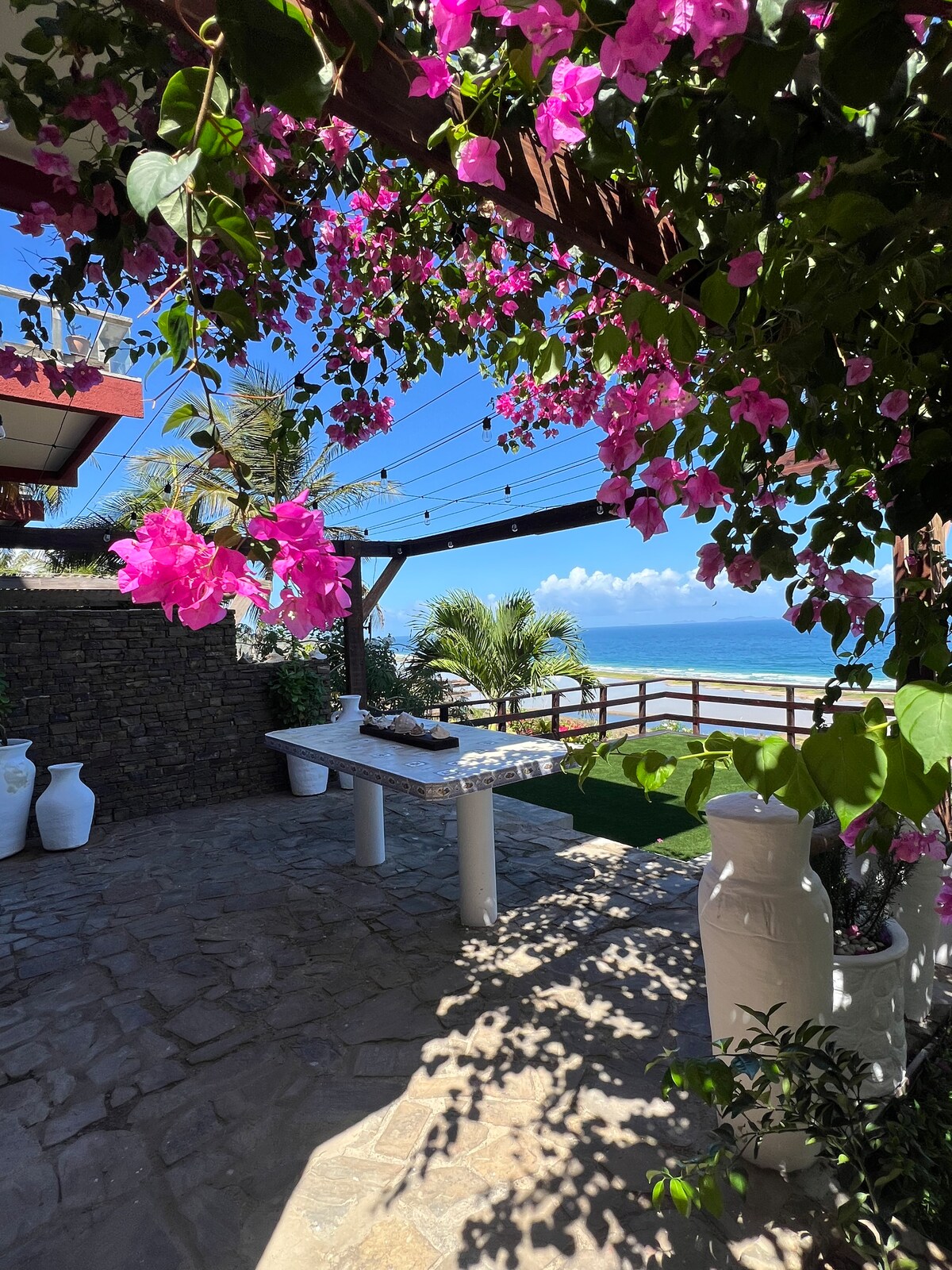 An outdoor patio is framed by vibrant pink bougainvillea, providing a scenic backdrop of the sea. A white stone table sits on the textured stone floor, surrounded by decorative vases. Lush greenery and a wooden railing enhance the tranquil coastal setting.