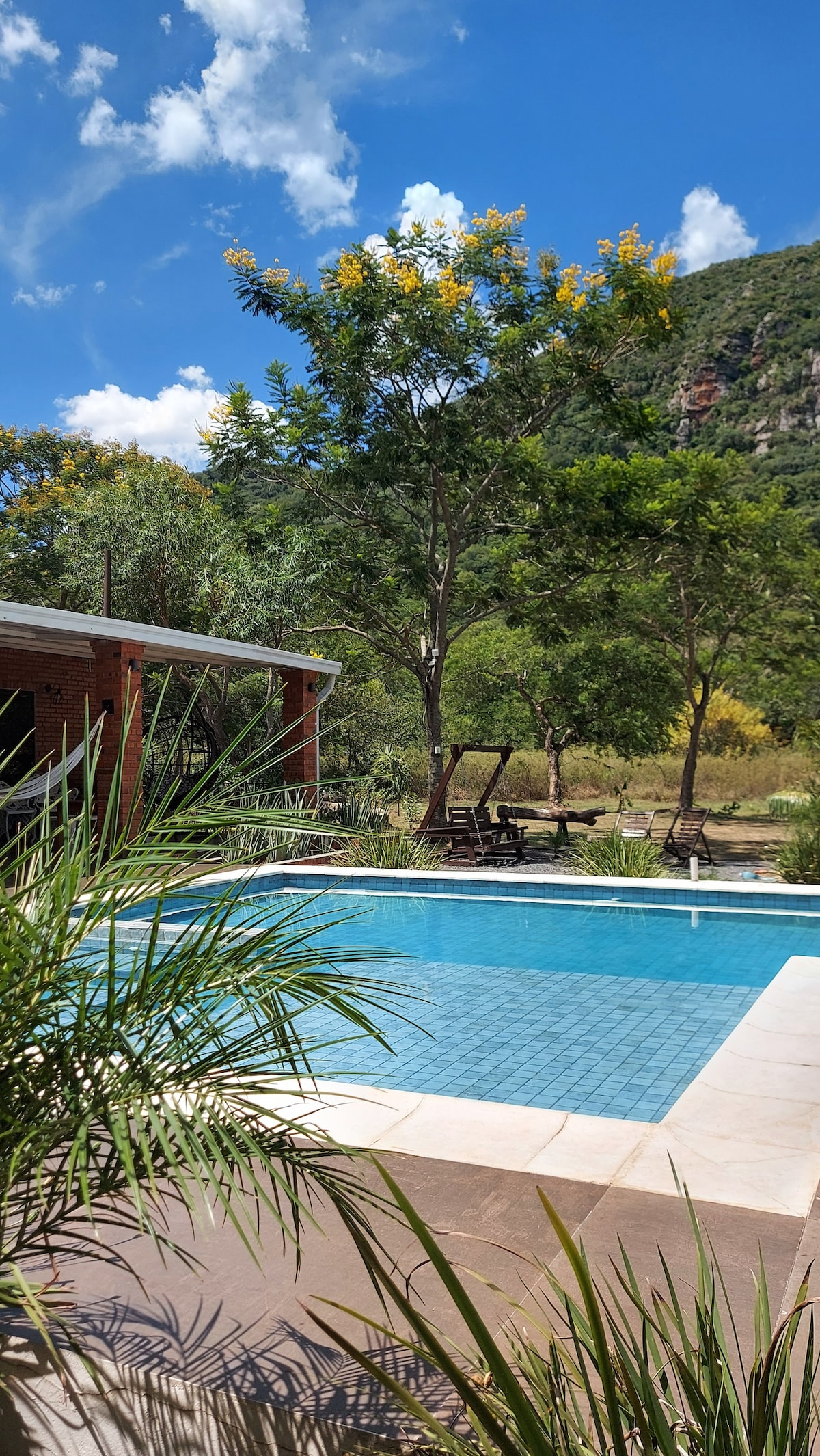 A clear blue pool is situated within a landscaped area, surrounded by lush greenery and trees. The backdrop features a hillside, enhancing the natural setting. Outdoor seating is provided nearby, allowing for relaxation under the open sky.