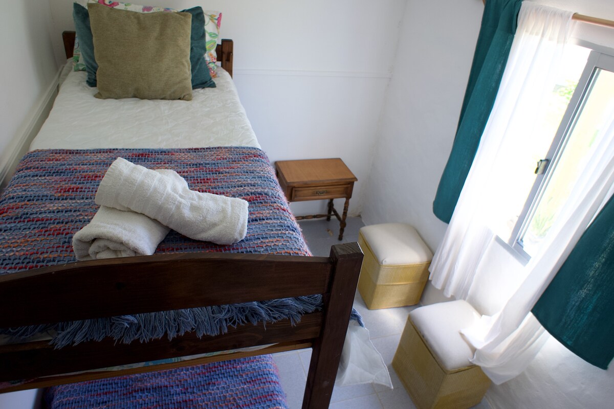 A private room features a single bed adorned with a textured blanket and neatly arranged towels. Nearby, a small wooden nightstand and two poufs provide additional comfort. Light filters through the window, accentuated by soft cream and turquoise curtains.