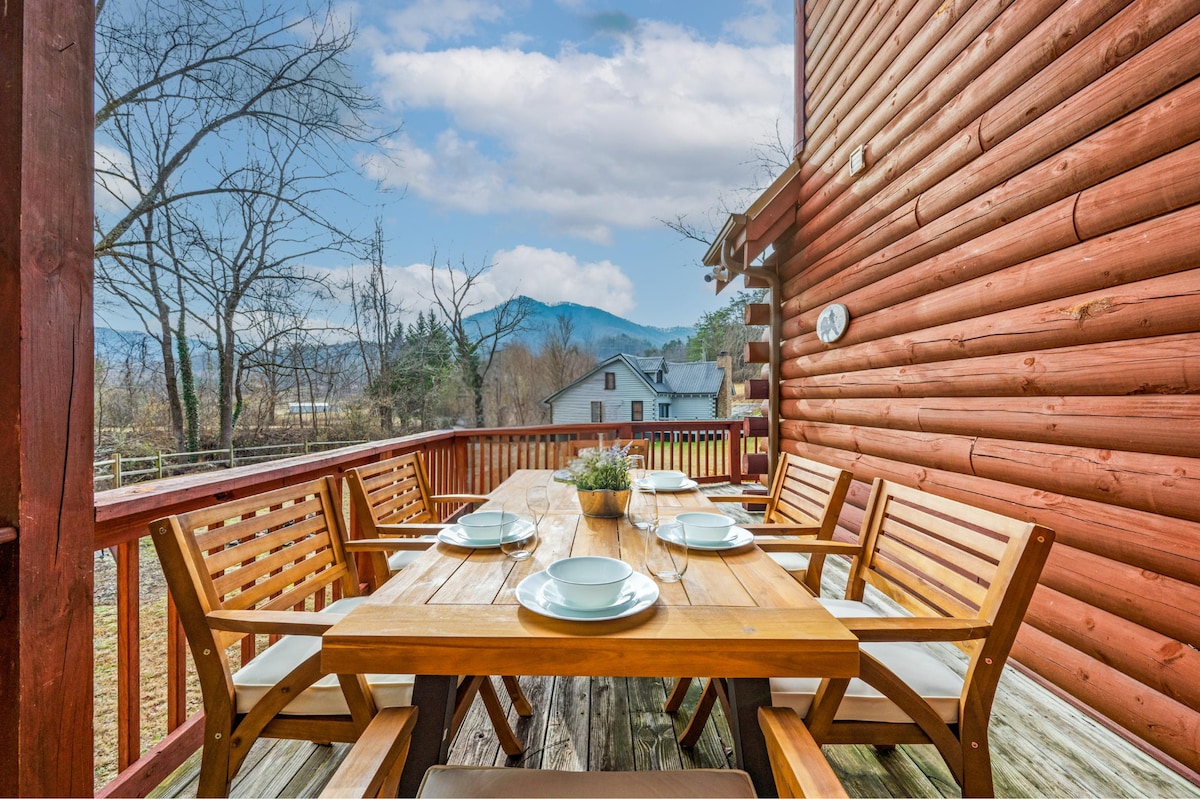 An outdoor dining area features a spacious wooden table surrounded by matching chairs, set against a backdrop of nearby mountains and trees. A serene atmosphere is created by the rustic log cabin exterior and the clear blue sky above.