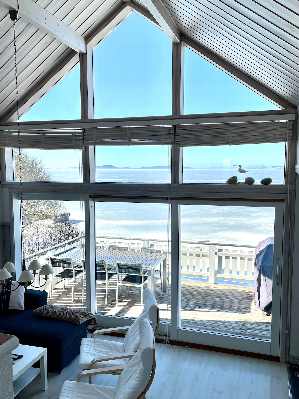 A spacious living area features a high ceiling with exposed beams, framed by large windows highlighting the ocean view. A comfortable seating arrangement is visible, alongside a dining table on the deck outside, inviting natural light into the room.