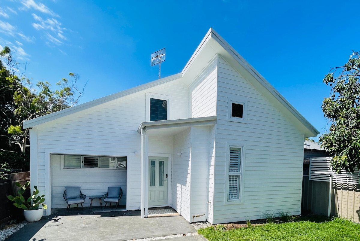 A modern guesthouse exterior showcases white cladding and a well-maintained lawn. Two seating chairs are positioned on the porch, complemented by a clear blue sky overhead. The entrance is framed by a small patio, enhancing accessibility and inviting entrance.