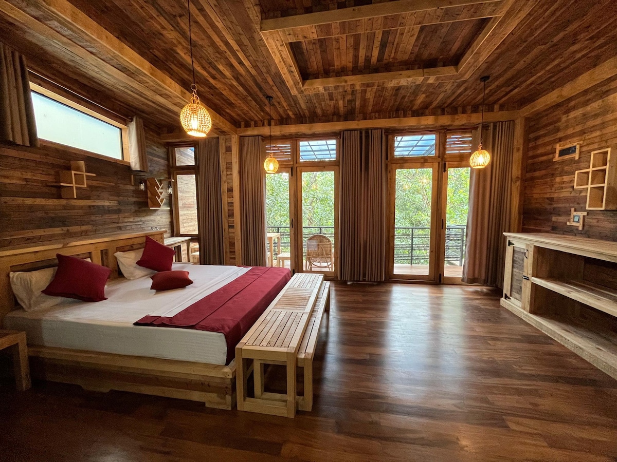 A spacious bedroom features reclaimed wood walls and ceiling, enhancing its natural charm. Large windows provide abundant natural light and offer views of greenery. A comfortable bed is complemented by a deep red runner, and wooden furniture adds to the room's warm aesthetic.
