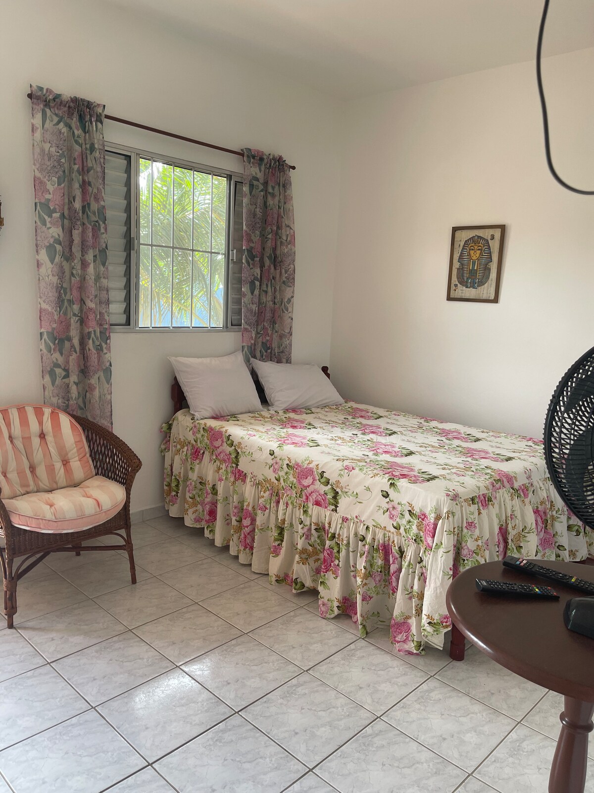 A cozy sleeping area is defined by a floral-patterned bedspread draping over a spacious bed, complemented by two white pillows. A small round table and a wicker chair enhance the space, while light filters through the curtains of the nearby window.
