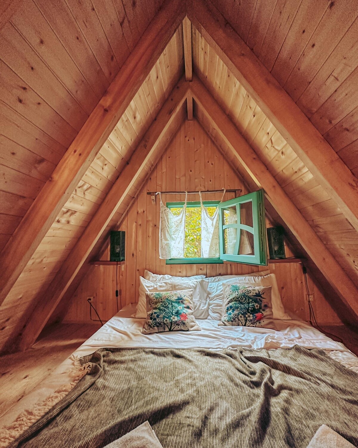 An A-frame sleeping area features wooden walls and a slanted ceiling, creating a cozy atmosphere. A spacious bed is adorned with decorative pillows and a soft blanket. Natural light enters through a green-framed window, partially dressed with light fabric curtains, providing a serene view of the outdoors.