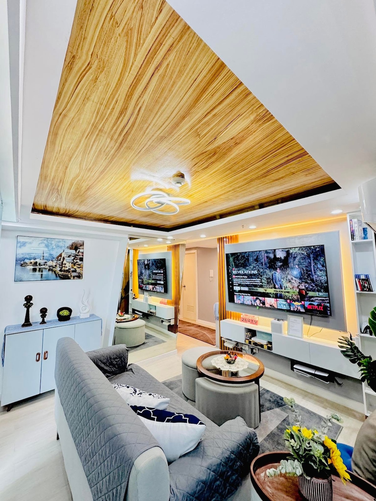A modern living room features a wooden ceiling design and a spacious sofa. A wall-mounted TV is positioned above a stylish console, while decorative accents, including flowers and artwork, enhance the space. Soft lighting creates a comfortable environment, suitable for relaxation and entertainment.
