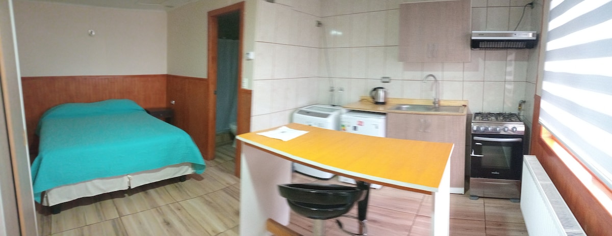 A cozy studio apartment is shown, featuring a spacious bed covered in a turquoise blanket. An adjacent kitchen area is equipped with essential appliances, including a stove and a washing machine. Natural light filters through a window, enhancing the warm tones of the wooden accents.
