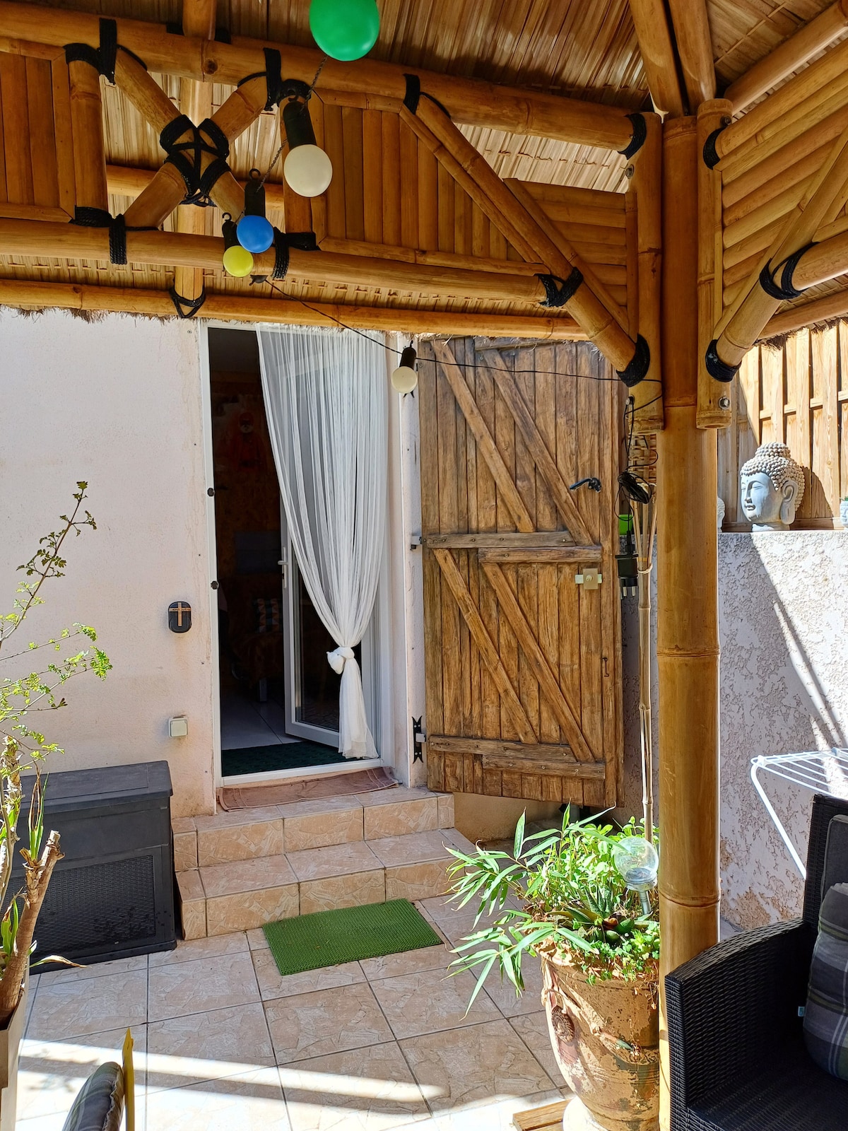 A bamboo gazebo shelters a welcoming outdoor space, featuring a doorway with white curtains leading into the studette. Decorative potted plants add a touch of greenery, while inviting seating is visible nearby. The area is well-lit, enhancing the inviting ambiance.