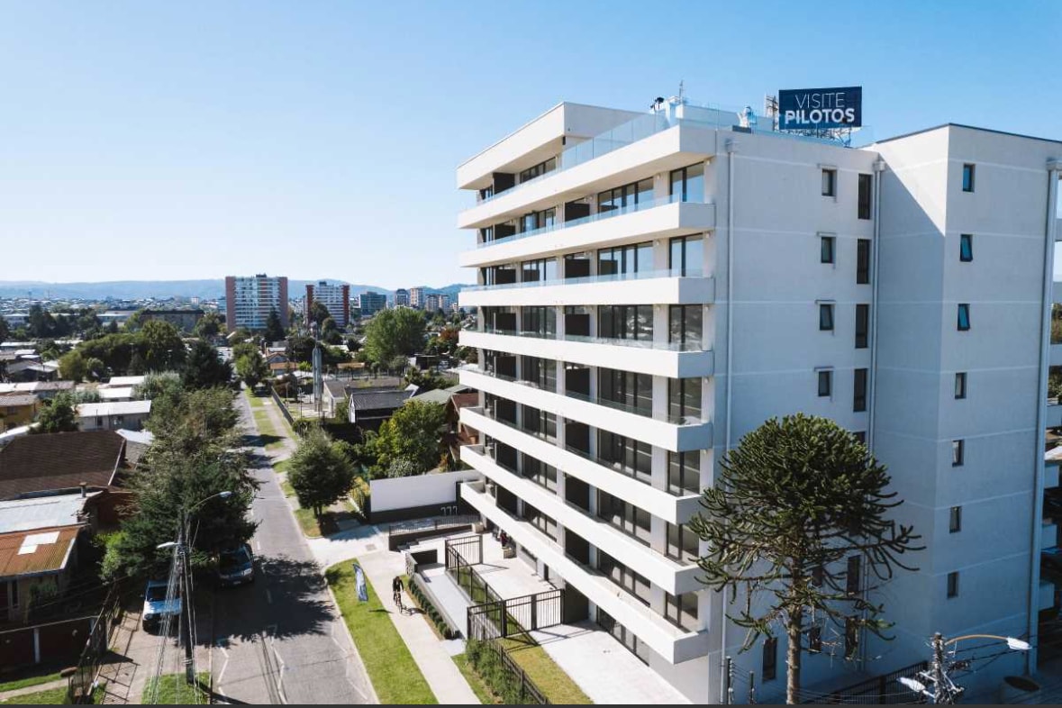 The exterior view of a modern building is presented, highlighting its five floors and clean lines. Surrounding green spaces are visible, along with nearby residential structures. The street in front features trees and power lines, contributing to the urban atmosphere.