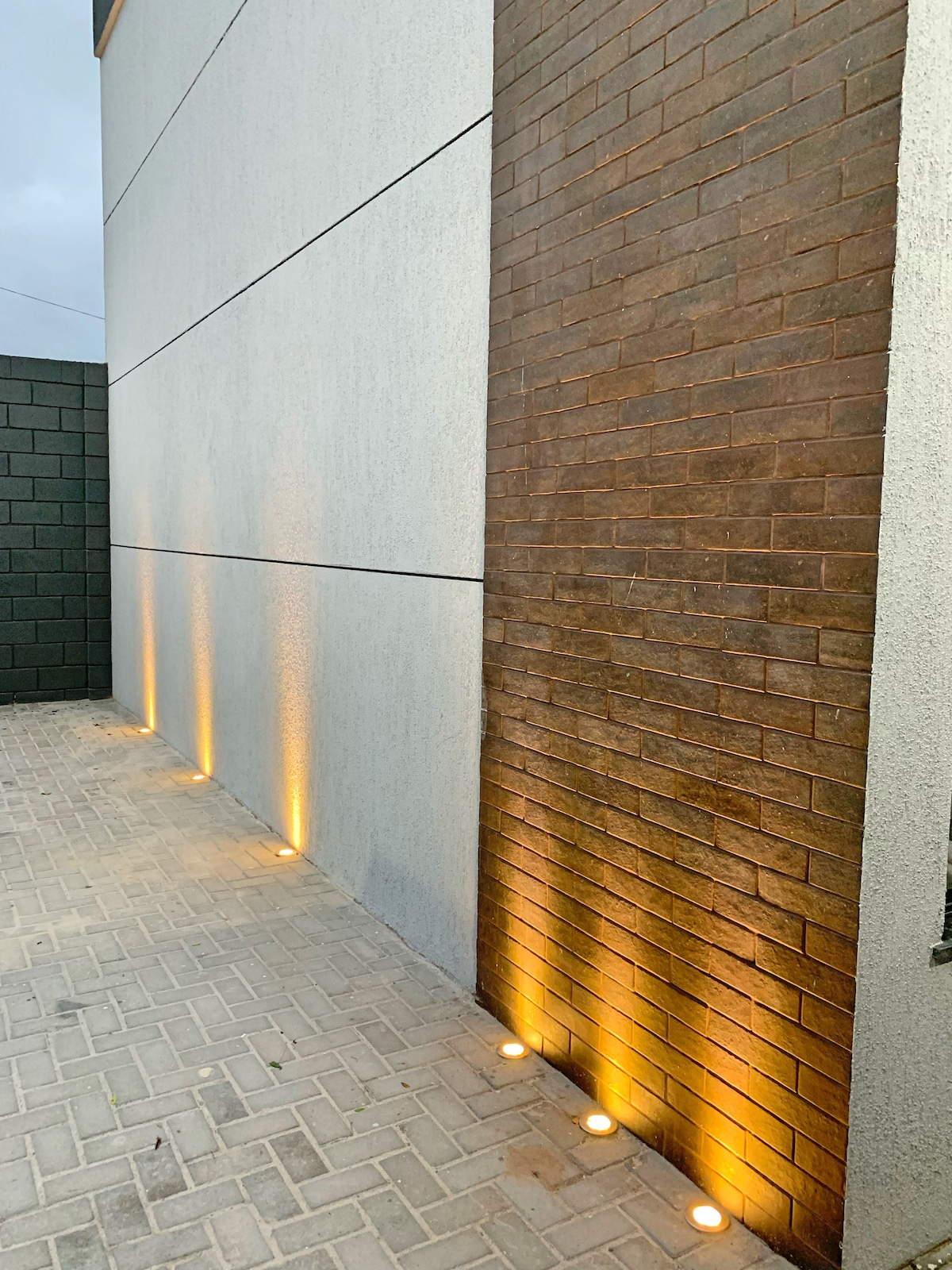 An exterior wall of the loft features a blend of textured gray and warm brown surfaces. Soft lighting is positioned along the ground, highlighting the brick and creating a welcoming ambiance. The paved area showcases neat stonework, contributing to the overall contemporary design.