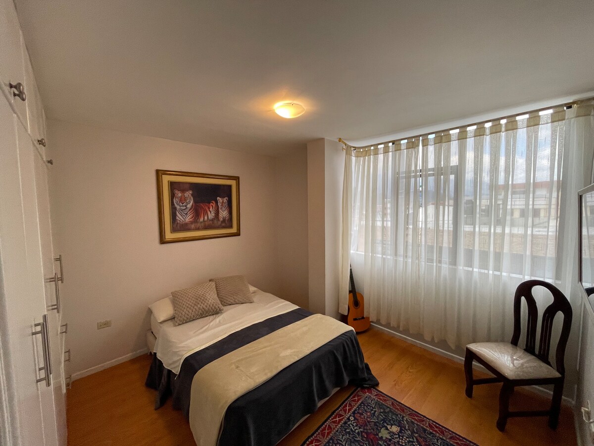 A well-lit bedroom featuring a spacious bed covered with a dark blanket and light-colored pillows. A framed artwork hangs above the bed, while a chair and an acoustic guitar are positioned nearby. Sheer curtains allow natural light to enter through the large windows.