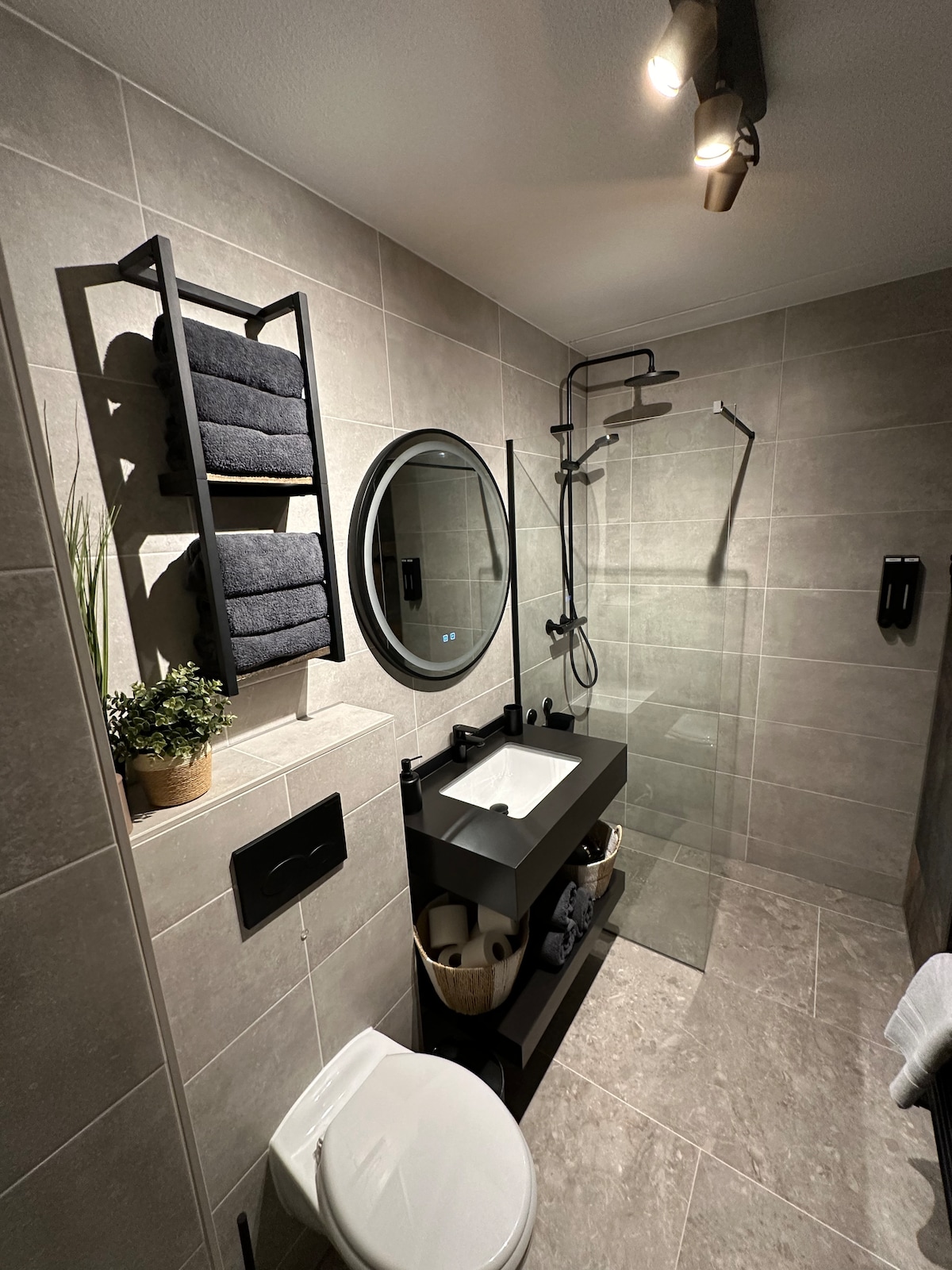 A contemporary bathroom features stylish gray tiles on the walls and floor. A circular mirror hangs above a modern sink, complemented by a black shelving unit holding neatly folded towels. A clear glass shower is visible, showcasing a sleek rain showerhead.