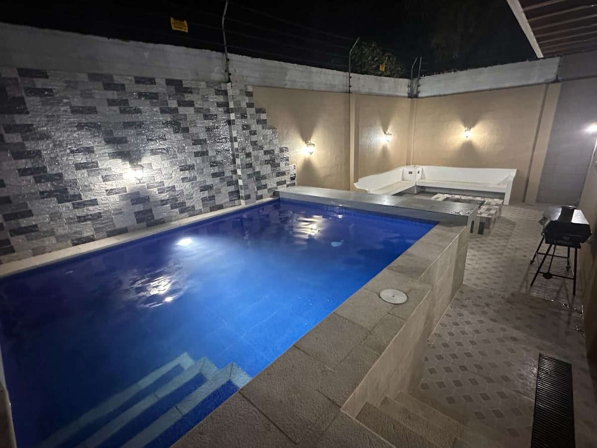 A tranquil swimming pool occupies the center of the outdoor space, surrounded by stone walls illuminated by soft lighting. A jacuzzi is positioned beside the pool, and a grill area offers additional amenities for outdoor gatherings.