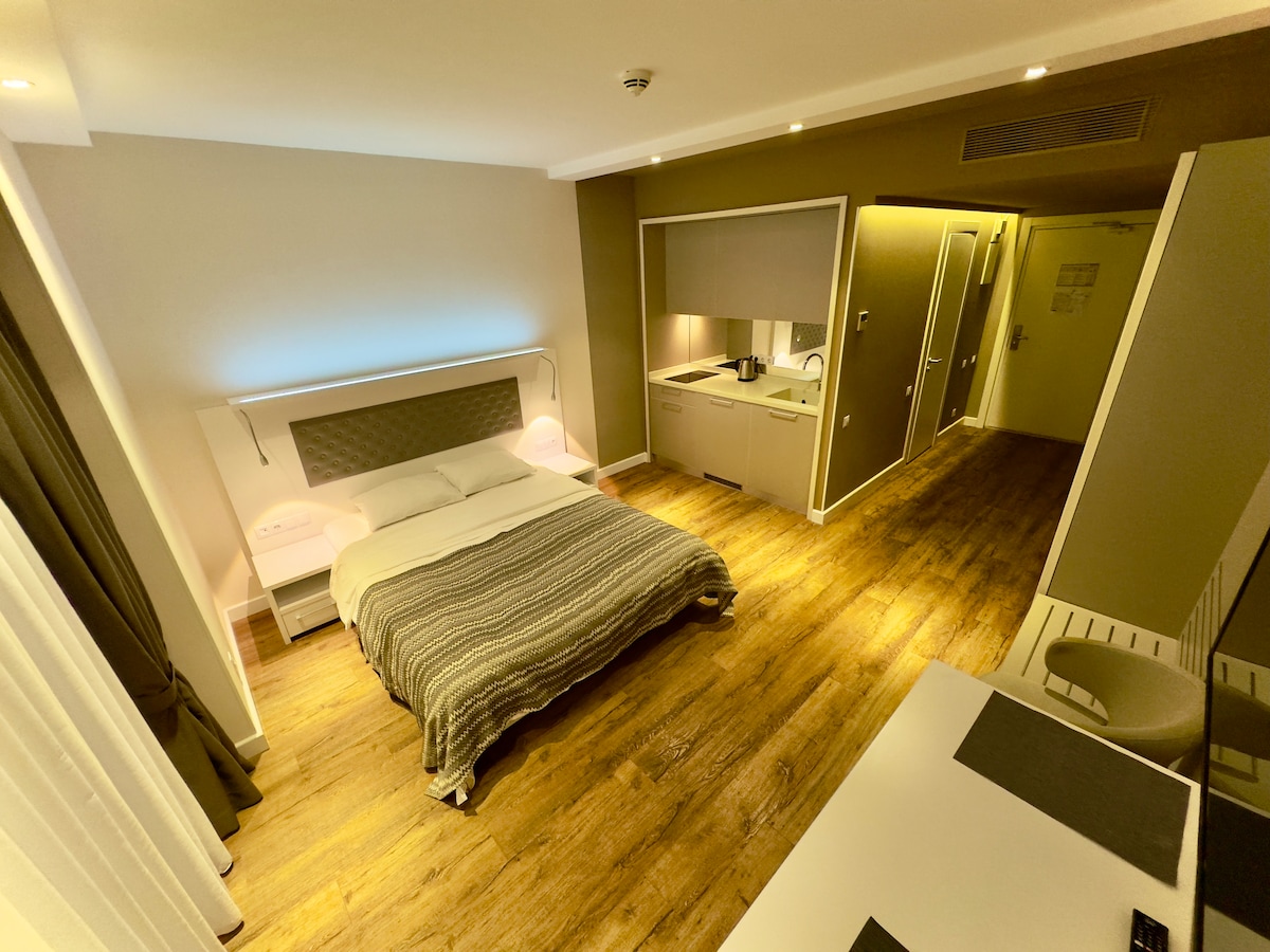 A modern studio room is shown, featuring a spacious bed with a textured gray blanket. Soft lighting illuminates the area, highlighting the adjacent kitchenette and dining setup. A neutral color palette is present throughout, complemented by warm wooden flooring.