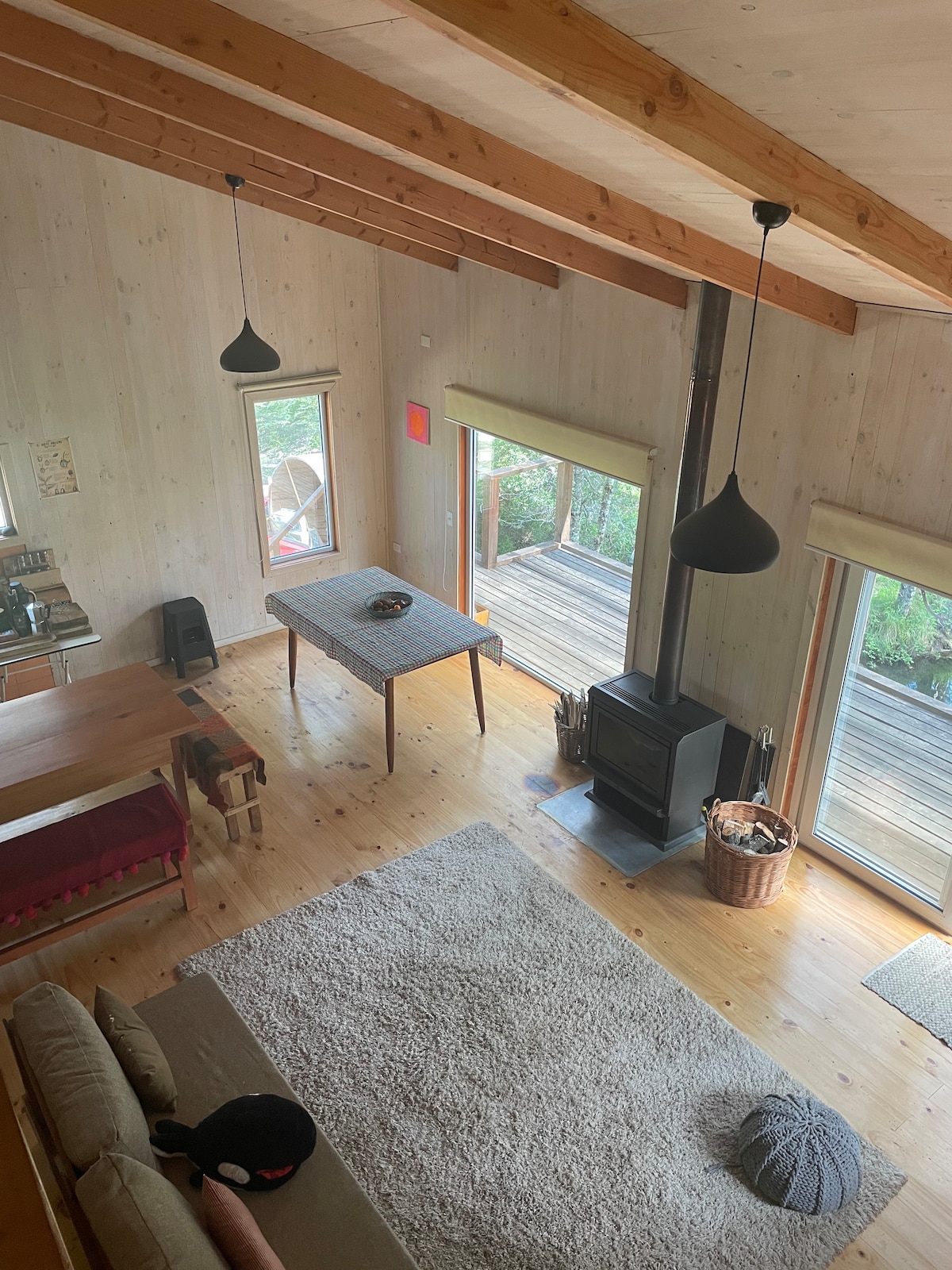 An aerial view of a cozy living space is presented, featuring wooden beams and walls. A comfortable sofa sits on a textured rug, while a dining table with a simple tablecloth occupies the center. Large windows offer natural light, and a wood-burning stove enhances the warmth of the setting.