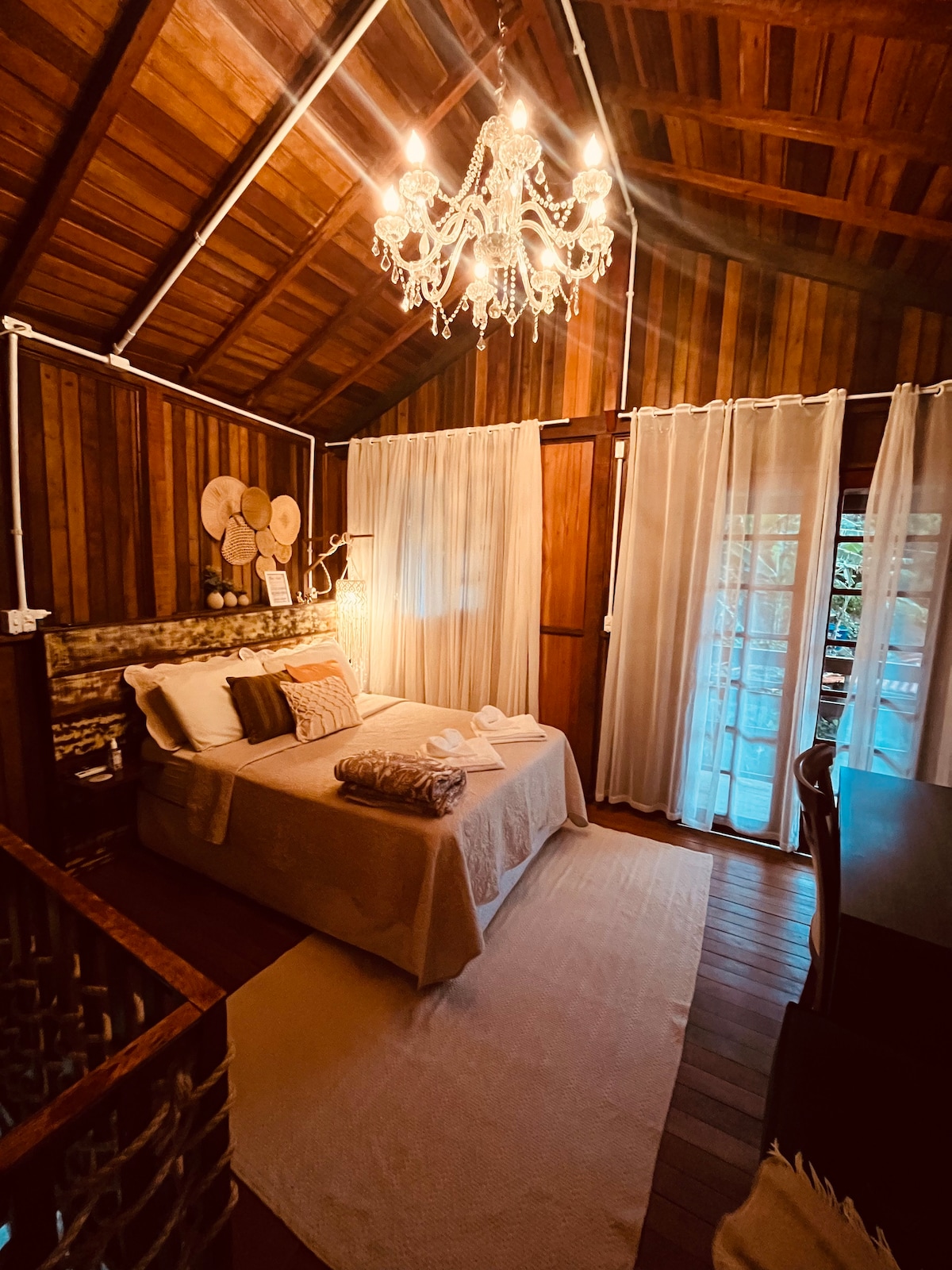 A warm wooden interior features a cozy sleeping area with a spacious bed, dressed in light linens and adorned with throw pillows. Soft lighting from a decorative chandelier highlights the textures of the wooden walls. Sheer curtains frame the window, adding natural light to the room.