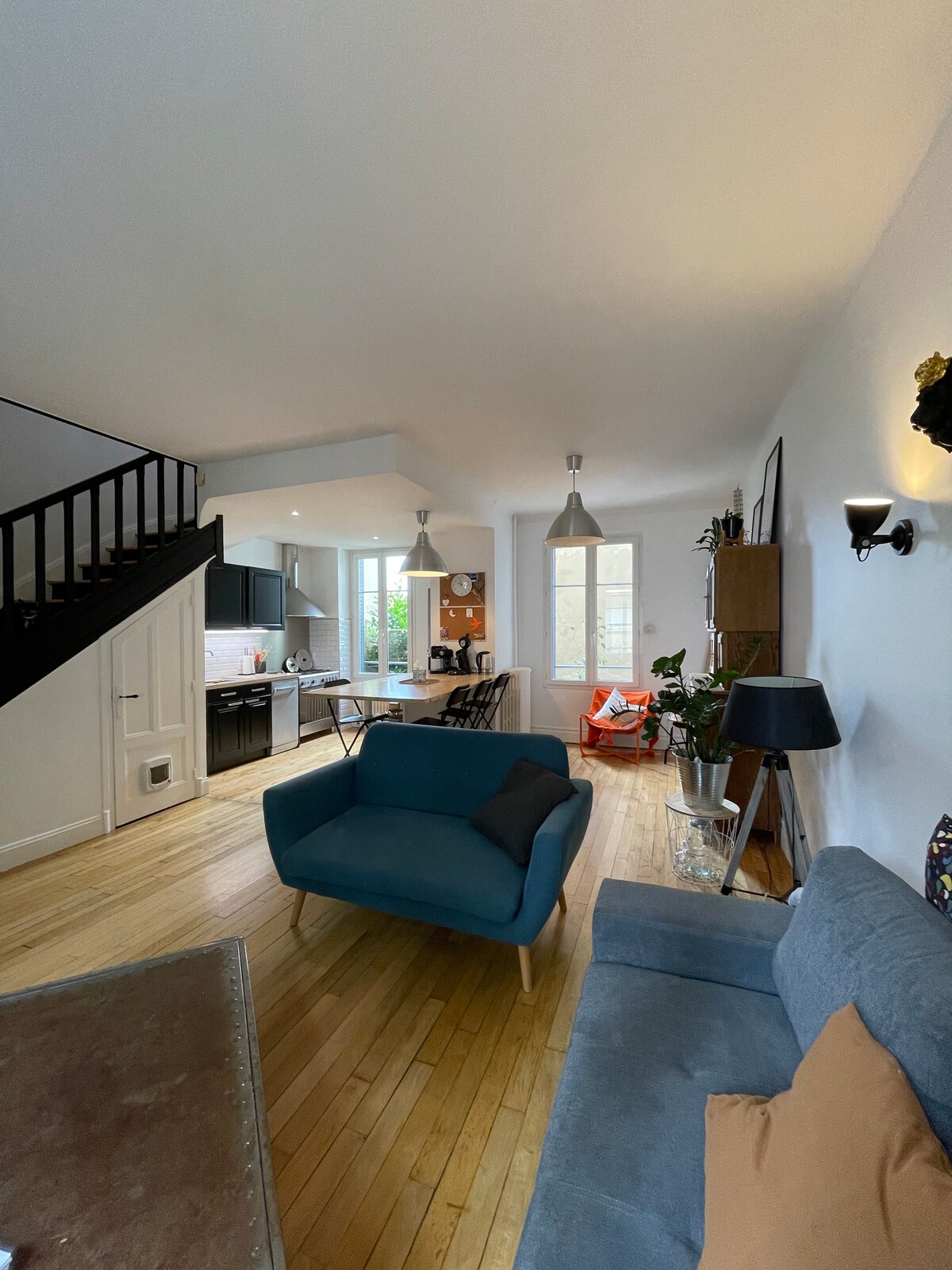 An open living space is observed, featuring wooden flooring and a light color palette. A blue armchair and grey sofa create a comfortable seating area. The kitchen area is visible, equipped with modern appliances. Large windows allow natural light to brighten the interior.