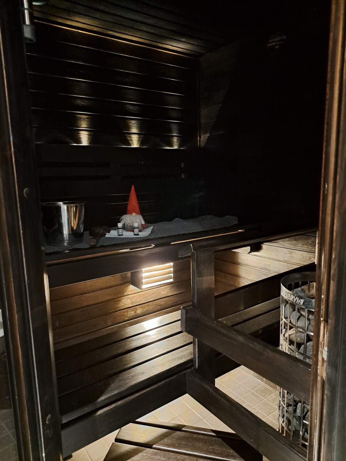 A wooden sauna is depicted, showcasing a serene interior with warm lighting. A small bucket and towel are positioned on a shelf. A waste bin and a decorative item are also visible, contributing to a tranquil environment. The walls are lined with dark wood, highlighting the peaceful setting.
