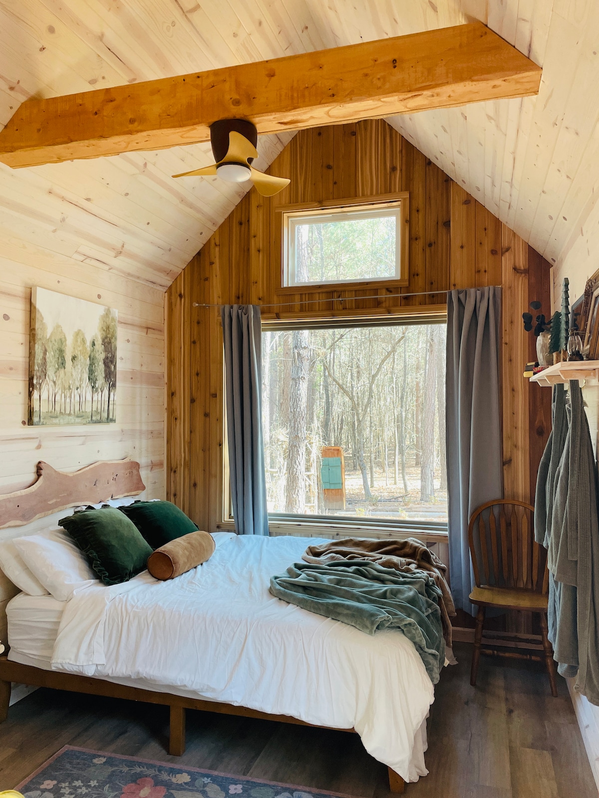 A warm wooden interior showcases a cozy sleeping area with a queen-sized bed adorned in soft linens and decorative pillows. Natural light filters through a large window, providing views of the surrounding forest. A chair and neatly draped towels complete the inviting space.