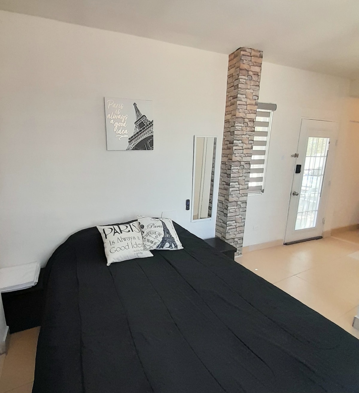 A spacious sleeping area features a black bedspread layered over a comfortable bed. A decorative pillow with a Paris-themed design adds a subtle touch. A minimalist mirror and a stone accent wall enhance the room’s modern look. A door leads to the exterior, allowing natural light.