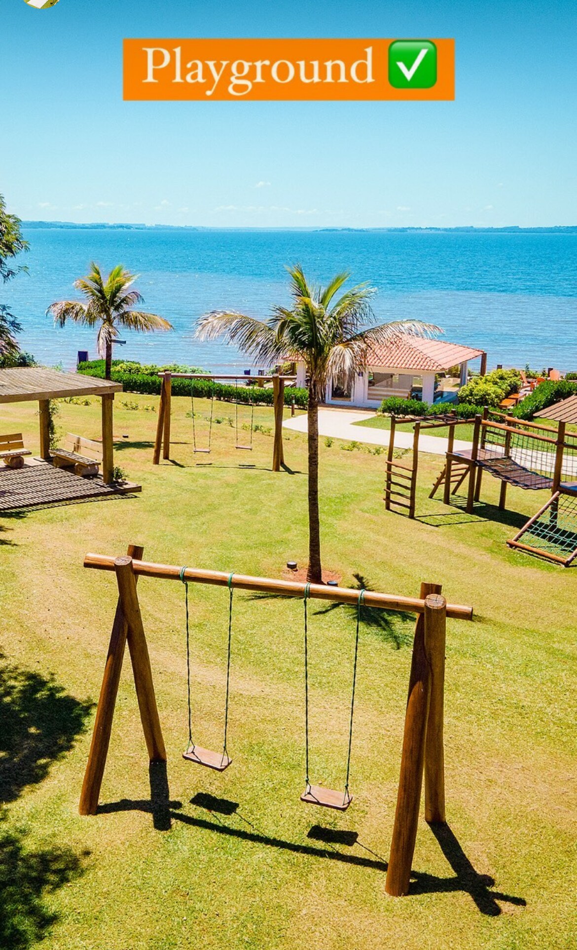 A grassy area features a wooden swing set framed by palm trees and overlooks the clean blue water. In the background, additional play structures and a house are visible, completing the inviting scene for outdoor activities.