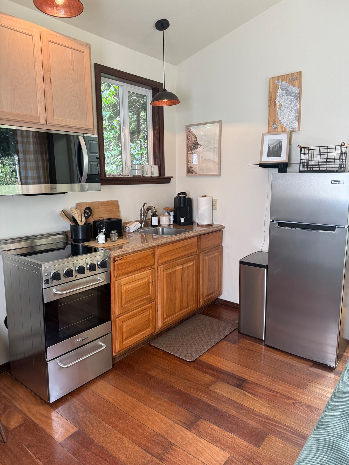 A well-equipped kitchenette features wooden cabinetry and stainless steel appliances. A gas stove, microwave, and mini fridge are visible, along with essentials like various utensils and a coffee maker. Natural light enters through a window, enhancing the warm tone of the wooden flooring.