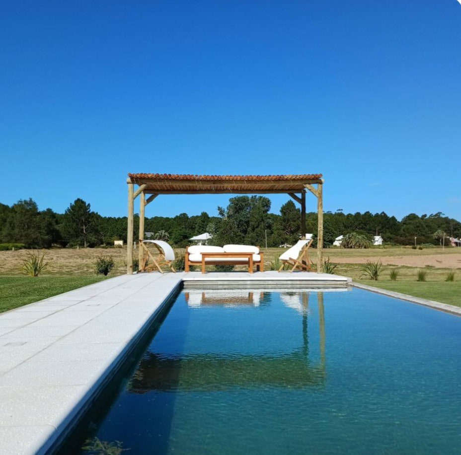 A refreshing pool is framed by a wooden pergola, providing shade over a seating area with a table and chairs. Sun loungers are positioned beside the clear water, surrounded by a manicured lawn that stretches toward natural greenery in the background.