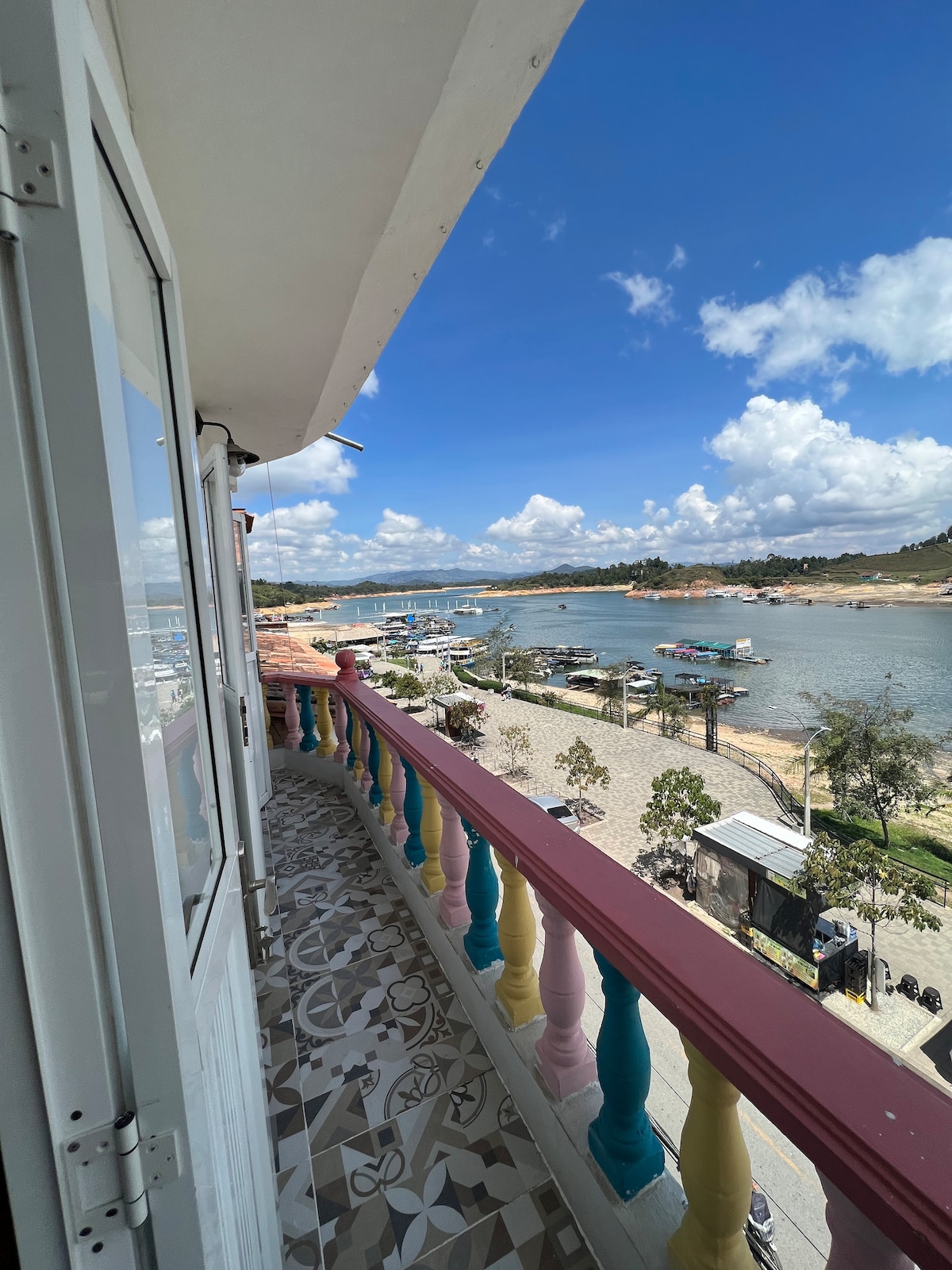 A balcony view showcases a vibrant scene of the Guatapé bay. Colorful railing frames the outdoor space, with boats visible on the water. The sky is clear with scattered clouds, creating a serene atmosphere. The tiled flooring adds a touch of design to the area.