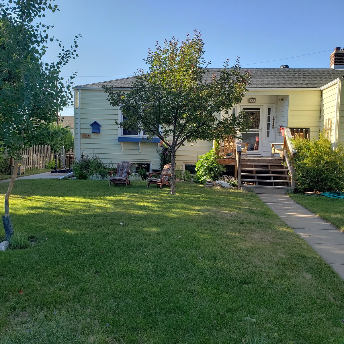 The exterior of a garden-level apartment is visible, showcasing a welcoming entrance with a ramp. Adjoining the entrance are two wooden chairs positioned on a grassy lawn, framed by a variety of foliage and a small tree. The space offers a relaxed atmosphere in a quiet neighborhood.