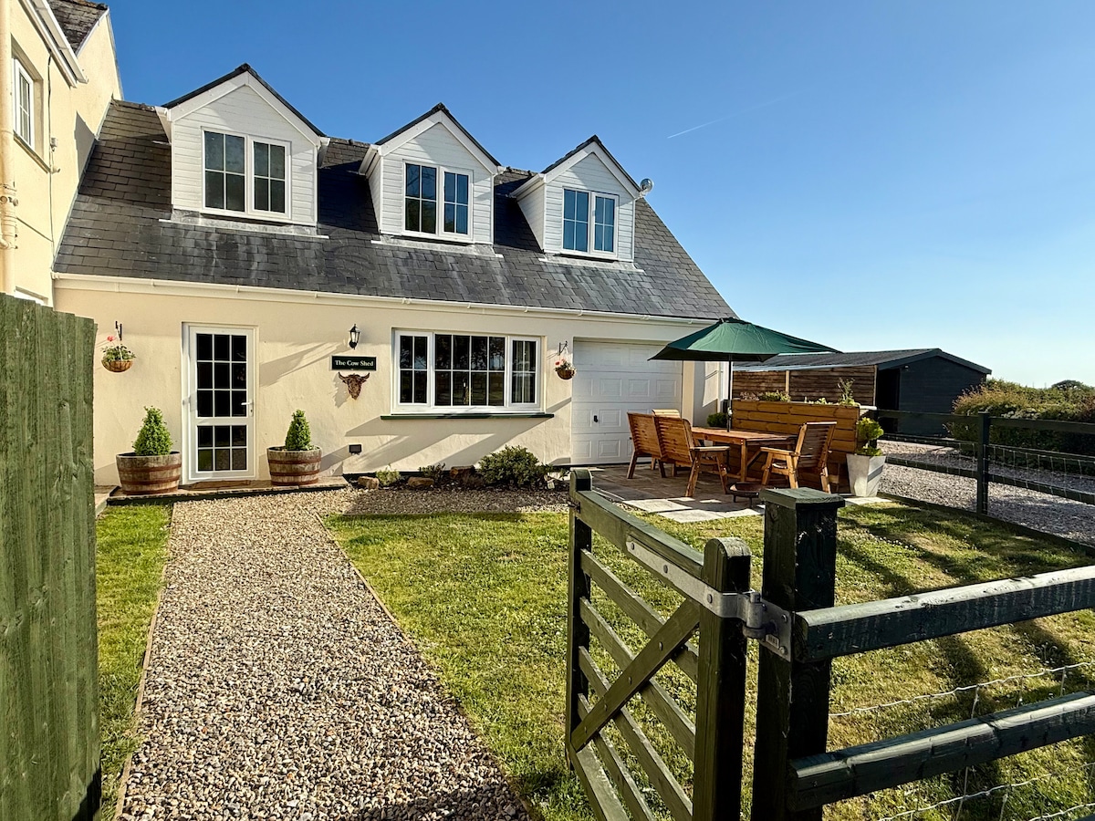 A charming two-story annex features a well-maintained front yard with a gravel pathway leading to the entrance. Outdoor seating is arranged under a green umbrella on a spacious wooden deck, providing an inviting space to relax. Near the house, a wooden shed is visible.