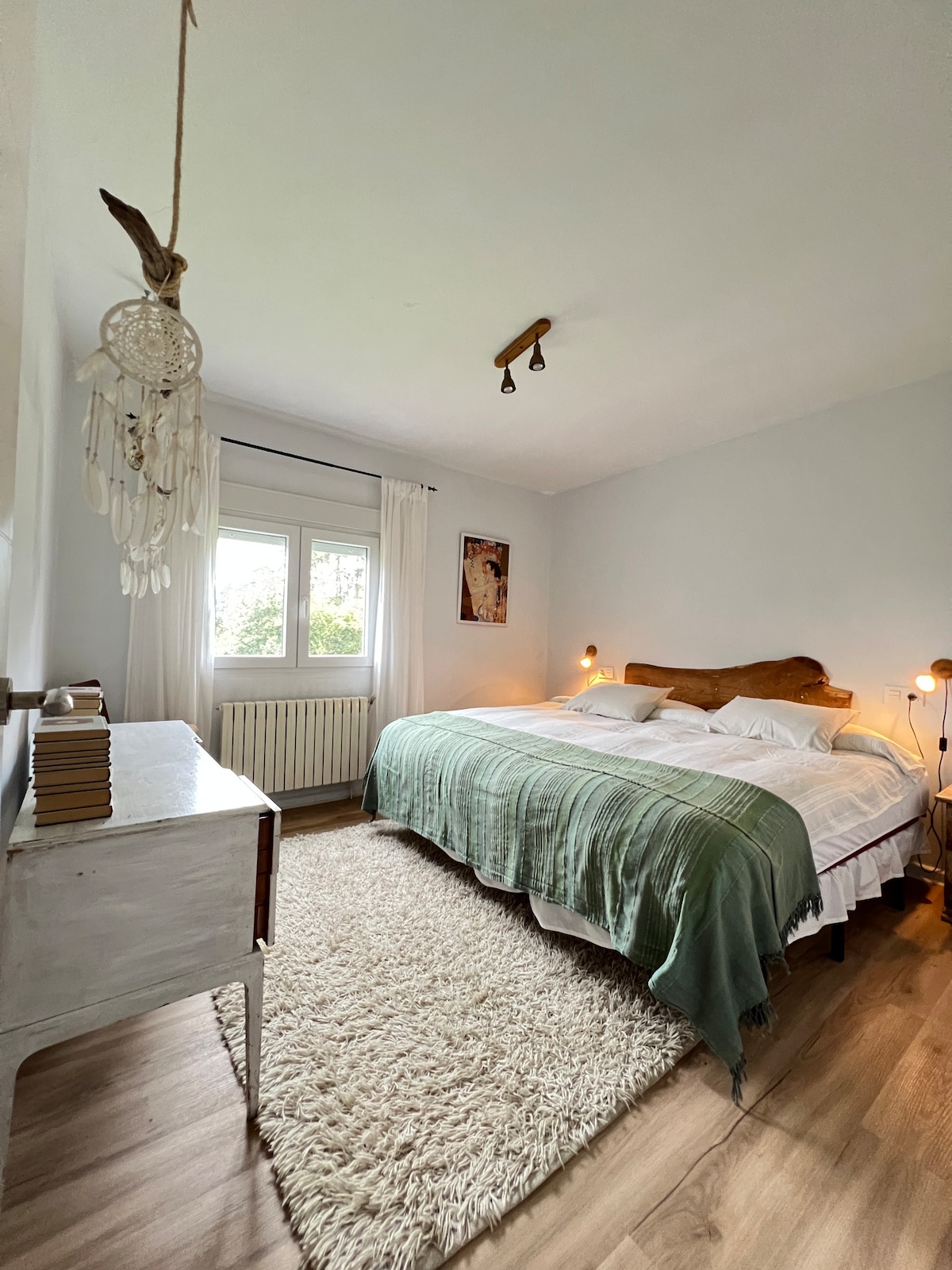 A spacious bedroom is presented with two cozy beds joined together for comfort. Soft, neutral linens are layered under a warm green throw. Natural light streams through a window, highlighting the serene decor which includes a light rug and a decorative piece on the wall.