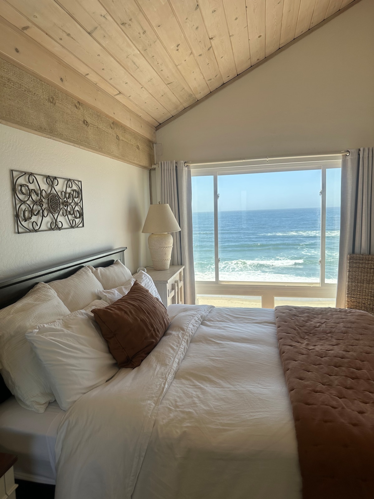 A serene bedroom features a spacious bed dressed in soft linens and a decorative throw. Natural light pours in through a large window, revealing sweeping ocean views and gentle waves. The room includes wooden accents, enhancing the coastal ambiance.