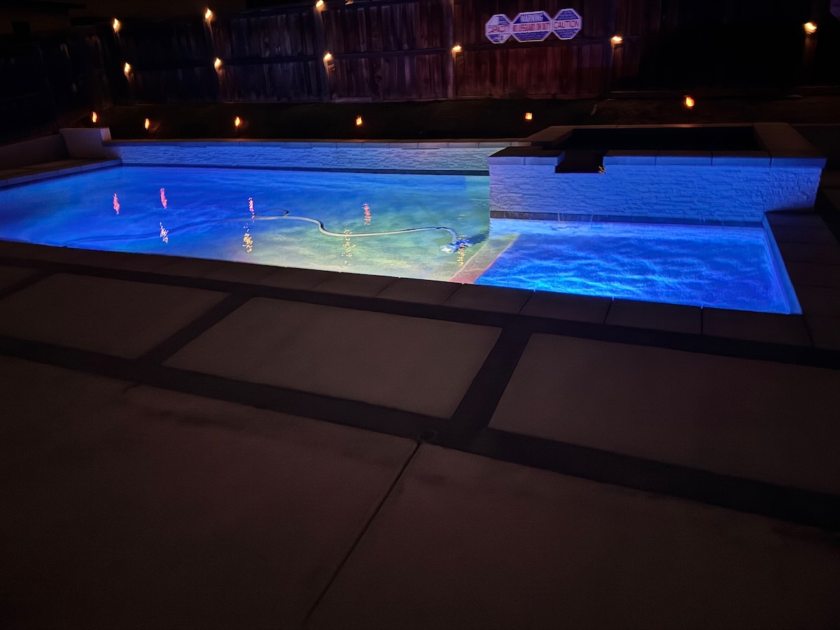 The image showcases a sparkling pool illuminated by vibrant lighting, creating a tranquil ambiance in the backyard. The water reflects a range of colors, complemented by the surrounding soft lighting along the edges. The pool's structured design features smooth stone accents and an inviting entrance.