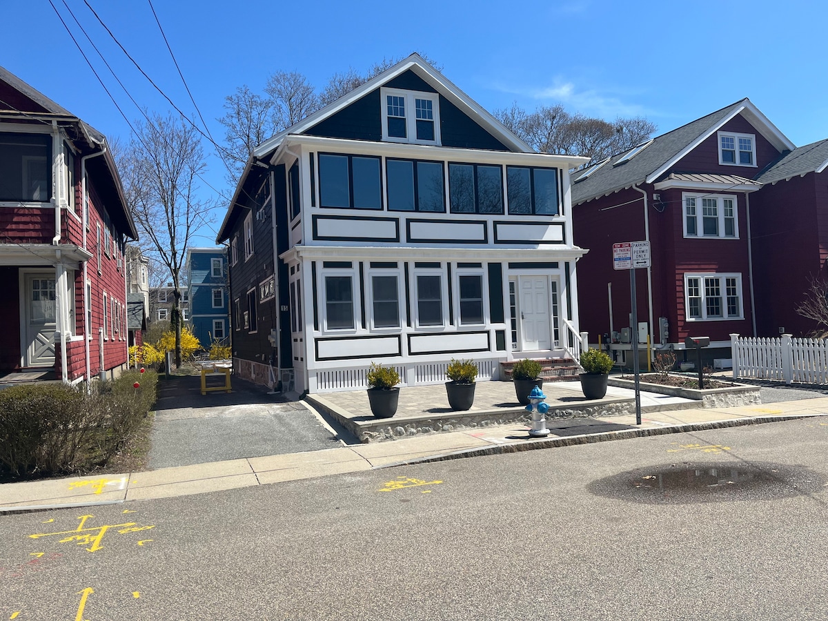 The exterior of a multi-story building is shown, featuring a mix of dark blue and white siding. A welcoming front porch with large windows is visible, and planters with greenery enhance the entry. A quiet street and neighboring red houses create a serene residential setting.