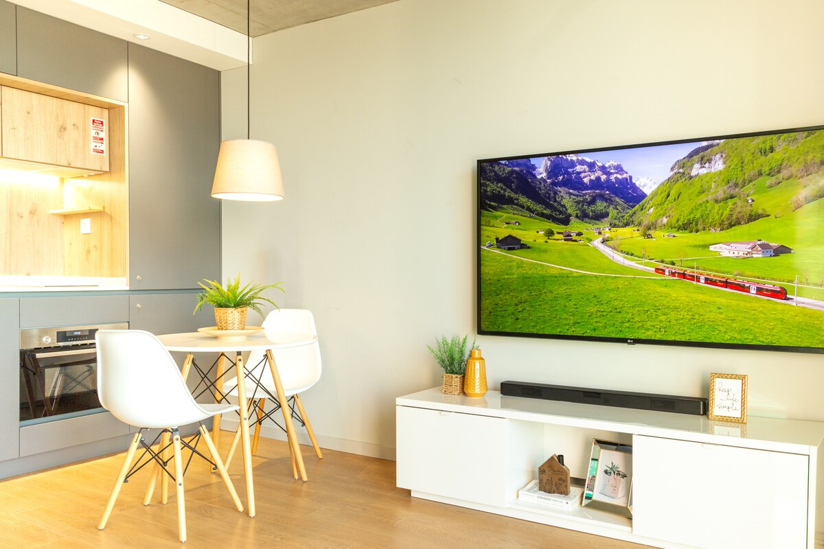A bright living area features a large wall-mounted TV displaying a scenic landscape. A compact dining table with white chairs sits adjacent to a stylish kitchen with wood accents. Modern decor and greenery add a fresh touch to the open space.