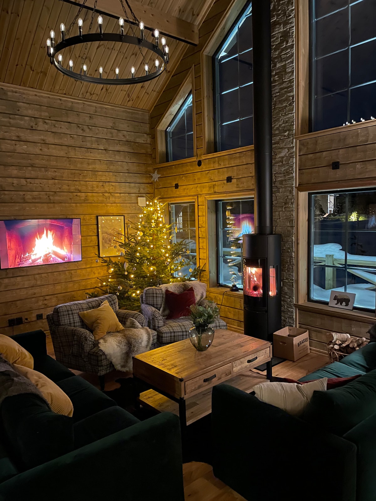 A cozy living area features large windows that provide views of the snowy exterior. A festive tree stands illuminated beside a comfortable seating arrangement. The dark wood and stone walls complement a modern fireplace, creating a welcoming atmosphere for relaxation.