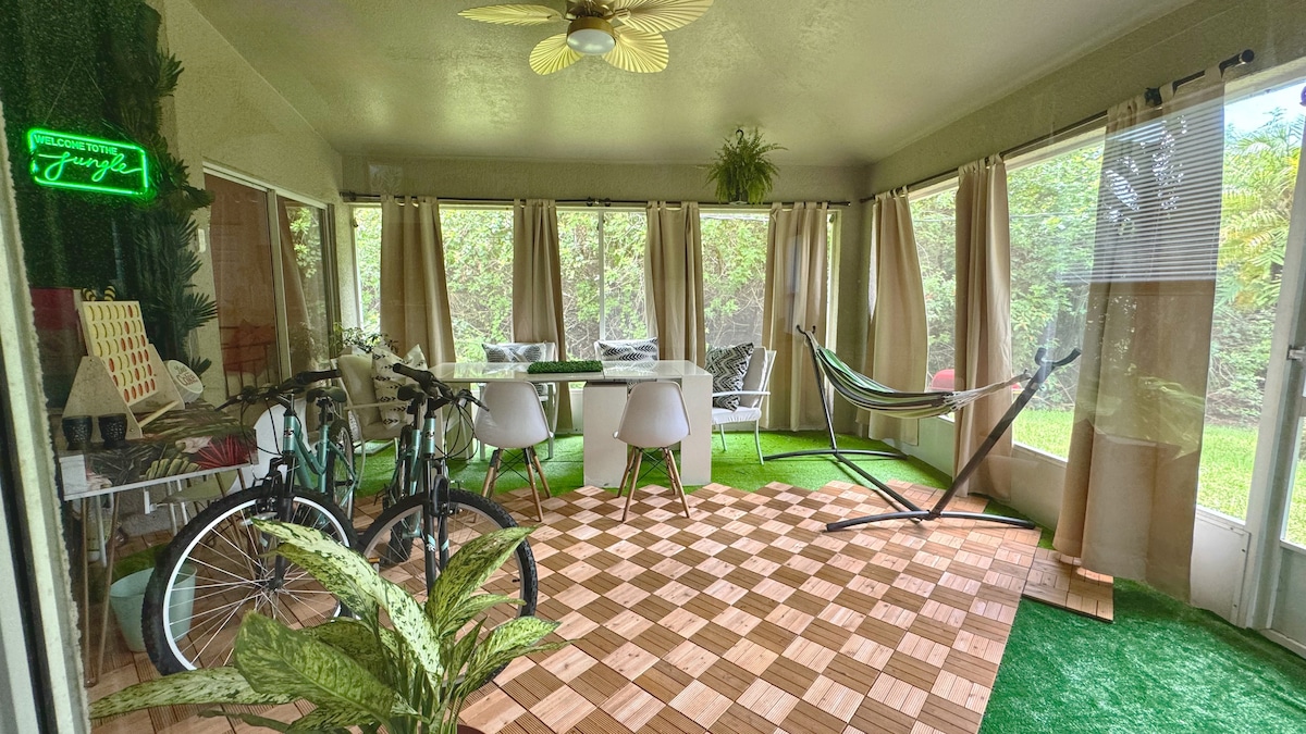 A bright Florida room is enveloped by large windows, revealing a green landscape outside. A dining table sits surrounded by white chairs, while two bicycles are positioned nearby. A hammock is comfortably hung, complementing the wooden flooring and indoor plants that enhance the cozy atmosphere.