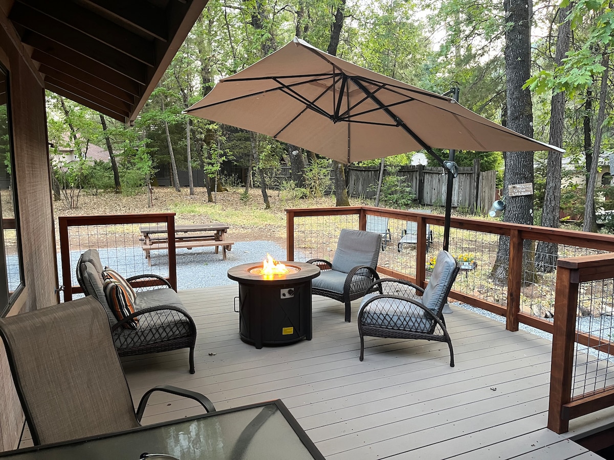 An outdoor deck area features a fire pit surrounded by four comfortable chairs. A large umbrella provides shade, and the wooden railing enhances the space's rustic charm. The surrounding wooded area adds a sense of privacy and tranquility.