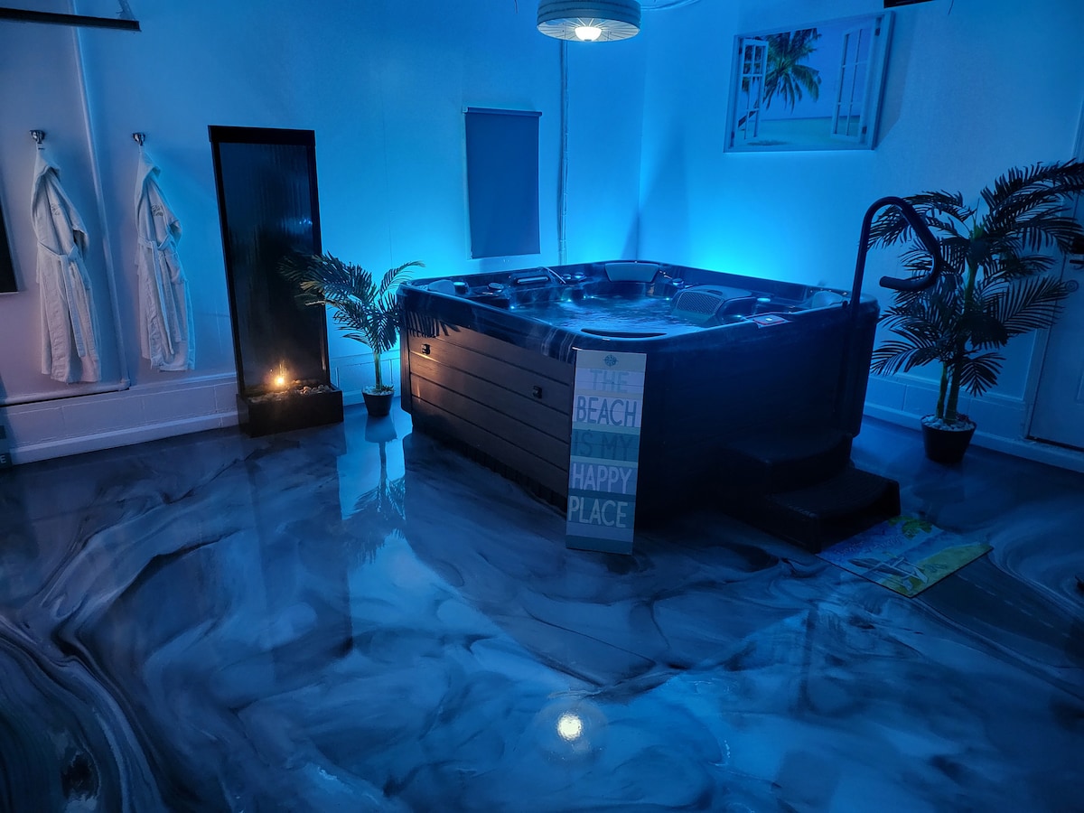 A hot tub is centrally placed in a softly lit space, surrounded by tropical-themed decorations. Calm blue lighting enhances the atmosphere, while large plants add a touch of nature. Soft robes hang on the wall, and a small candle casts a gentle glow nearby.