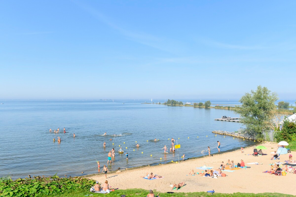 A sandy beach extends along the shoreline, with sunbathers relaxing on towels. Gentle waves lap at the edge, while various individuals enjoy swimming and playing in the water. Sun umbrellas provide shade in select areas, and a clear blue sky stretches above the scene.