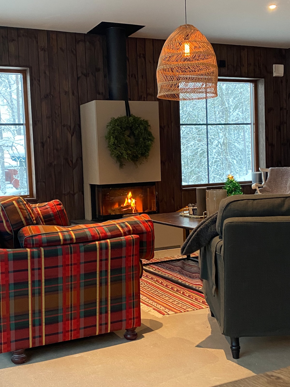 A cozy sitting area features two armchairs, one with a plaid pattern and the other in a solid color, positioned near a modern fireplace. A woven pendant light hangs above, while individual decor pieces add a touch of warmth. Snow can be seen through the large windows.