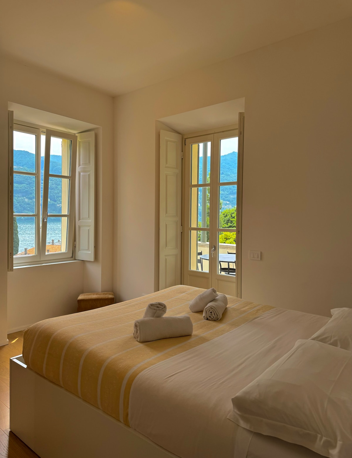 A bright bedroom features large windows framing scenic views of the lake and surrounding mountains. The spacious bed is centered in the room, adorned with rolled towels. Light-colored walls enhance the airy atmosphere, while natural light brightens the space.
