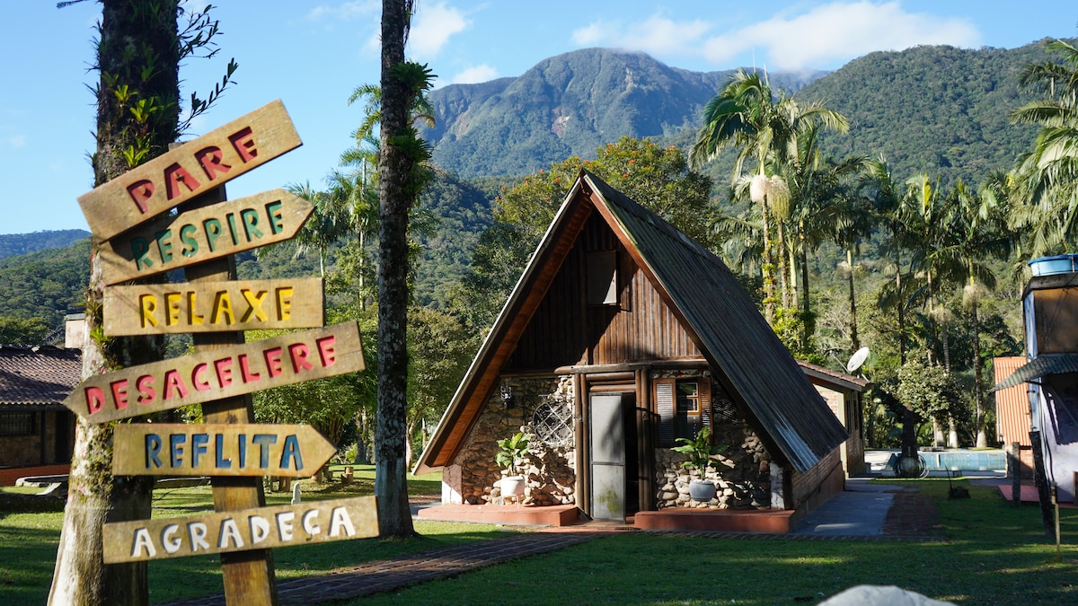 A charming wooden chalet stands against the backdrop of a green mountain, surrounded by palm trees. Colorful directional signs are planted in the manicured lawn, offering various suggestions for relaxation and mindfulness. The clear blue sky enhances the tranquil setting.