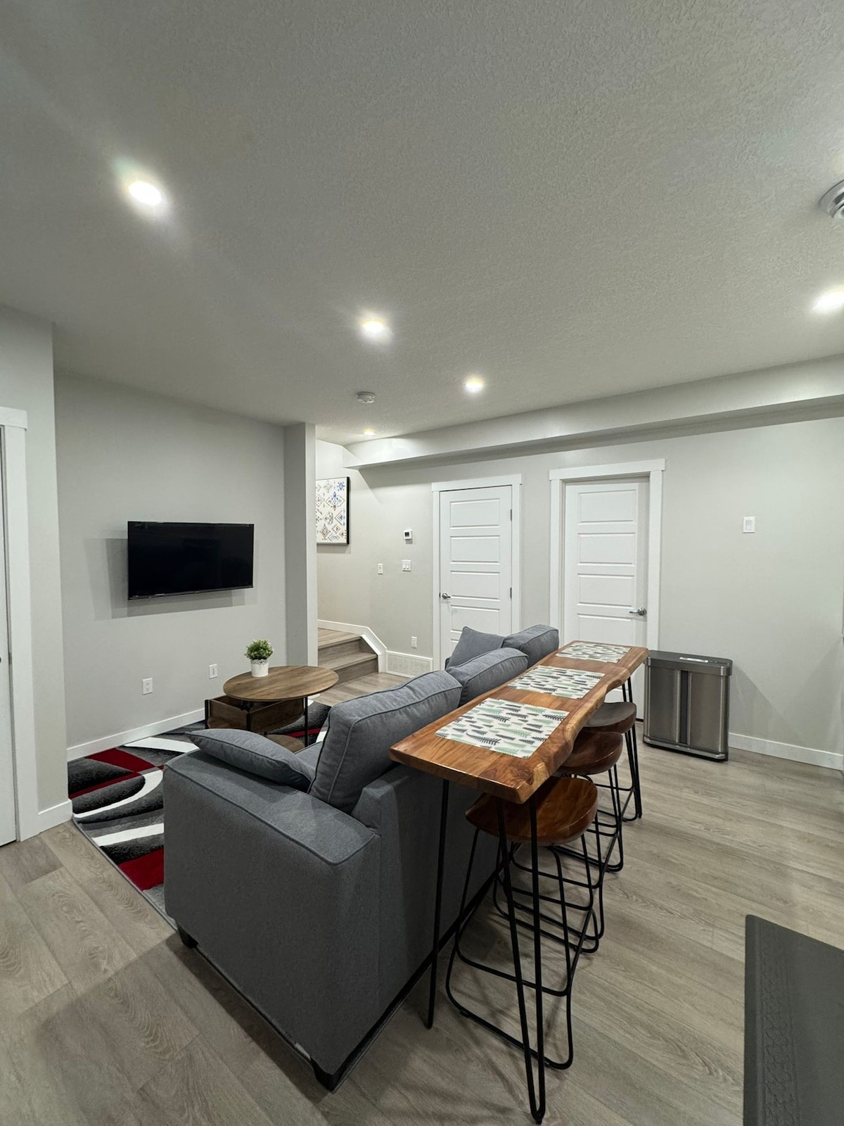 A contemporary living area features a gray sectional sofa facing a mounted flat-screen TV. A wooden dining table sits nearby with high stools, while two white doors offer access to separate rooms. Underfoot, light-colored flooring complements the modern design.