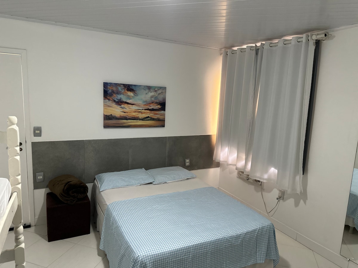 A bright bedroom features a spacious bed dressed in blue checkered linens, with two pillows on top. Light streams in through sheer white curtains, and a painting of a serene landscape adorns the wall above the bed. A small wooden nightstand is positioned beside the bed.