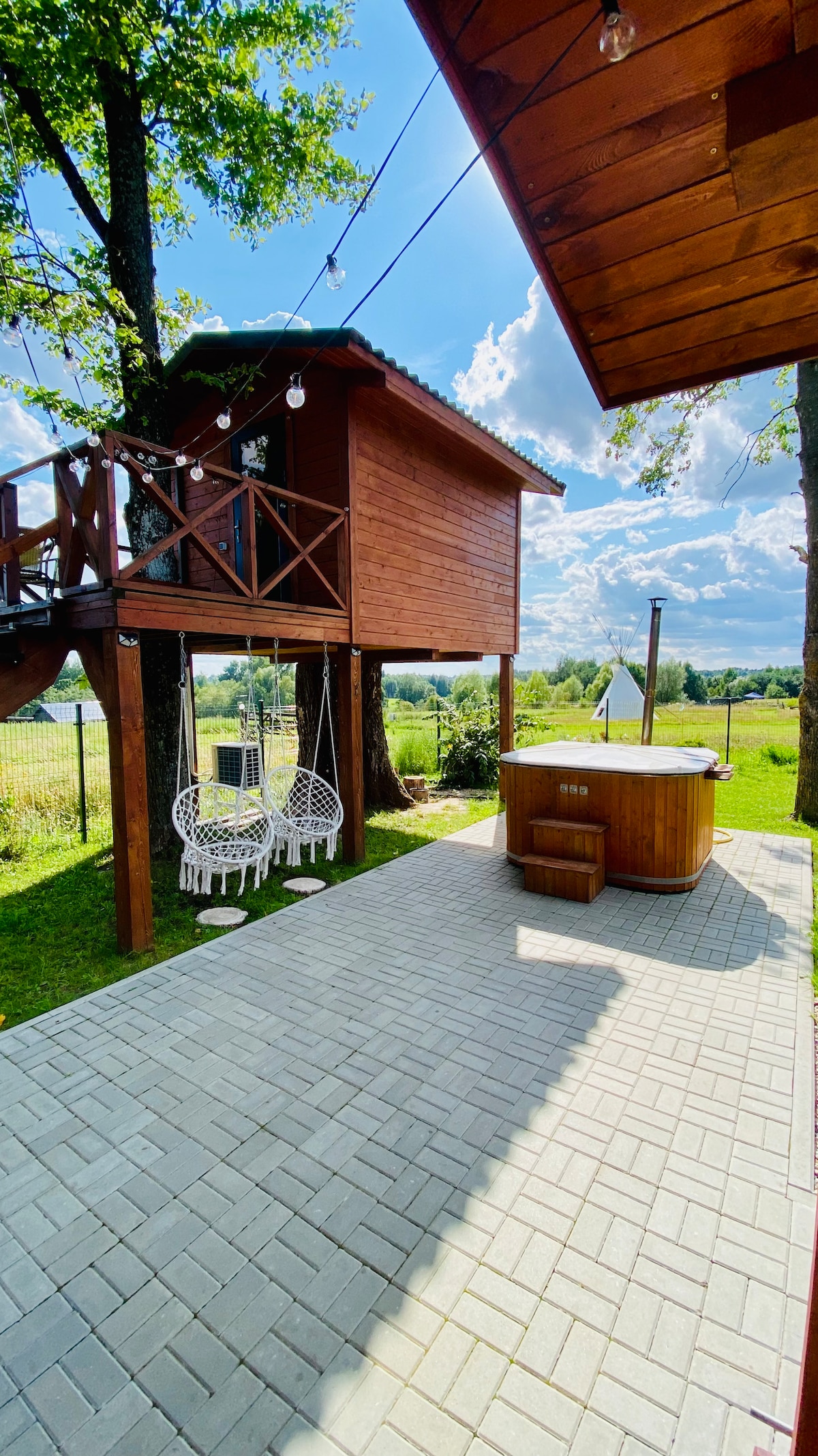 A wooden treehouse is elevated above the ground, surrounded by greenery. Below, two hanging chairs are suspended from a nearby tree. A hot tub is positioned on a spacious patio paved with light-colored stones, providing an inviting outdoor space.