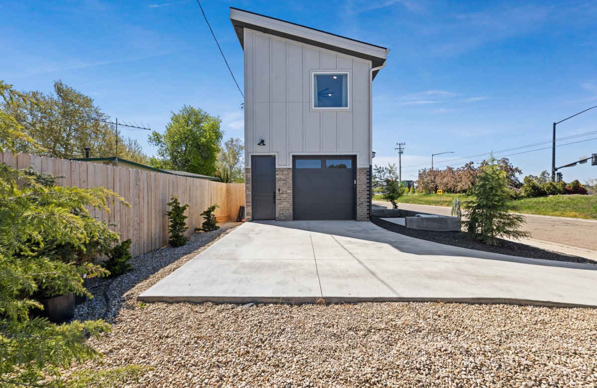 The exterior of a modern dwelling features a light-colored façade with stone accents. A concrete driveway leads to a garage door. The landscape includes gravel and small shrubs, with a fence bordering the property. Large windows are visible on the upper level.