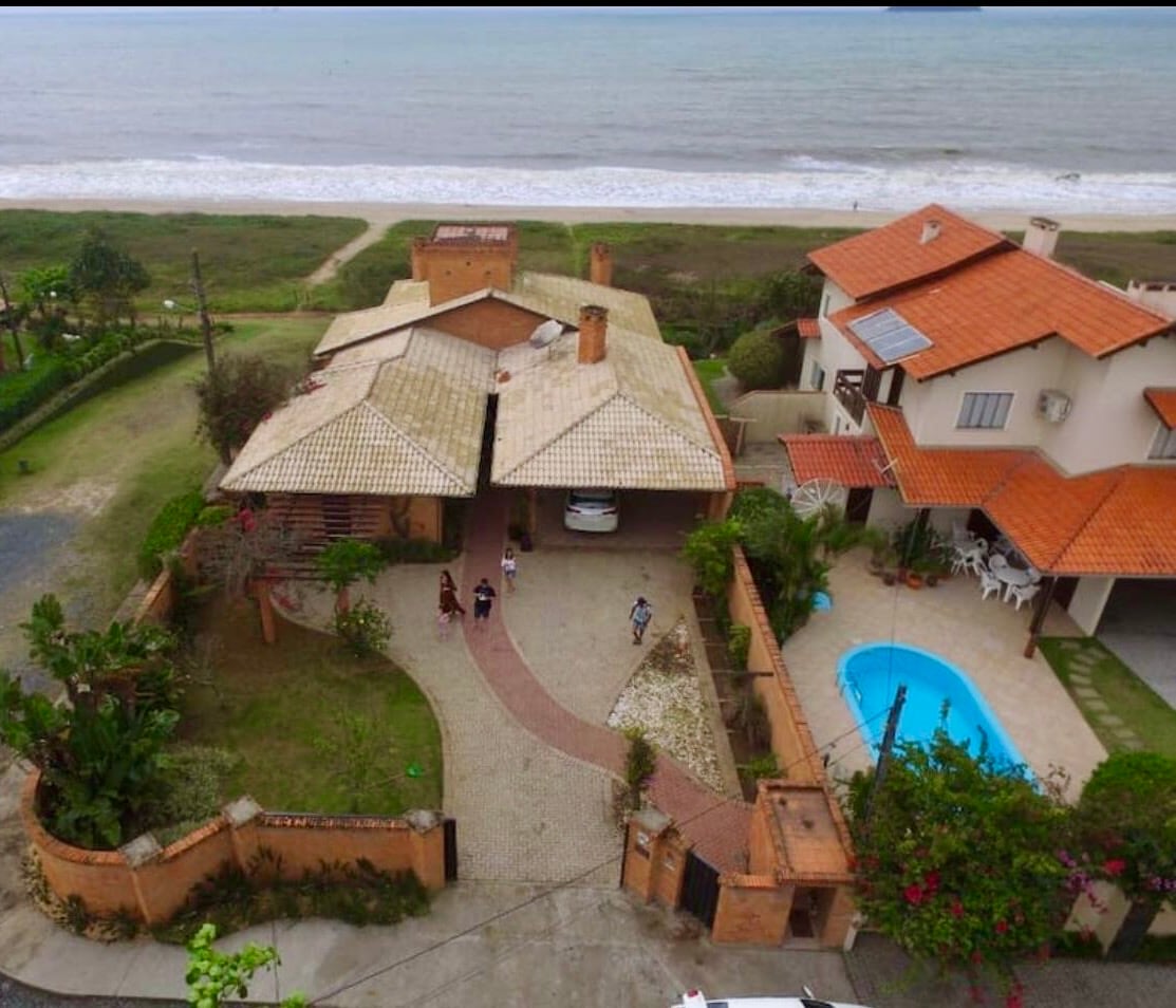 An aerial view captures a beachside property featuring two distinct structures with red-tiled roofs. A clear swimming pool is visible beside a paved driveway that leads to the entrance. Lush greenery surrounds the area, with the ocean visible in the background.