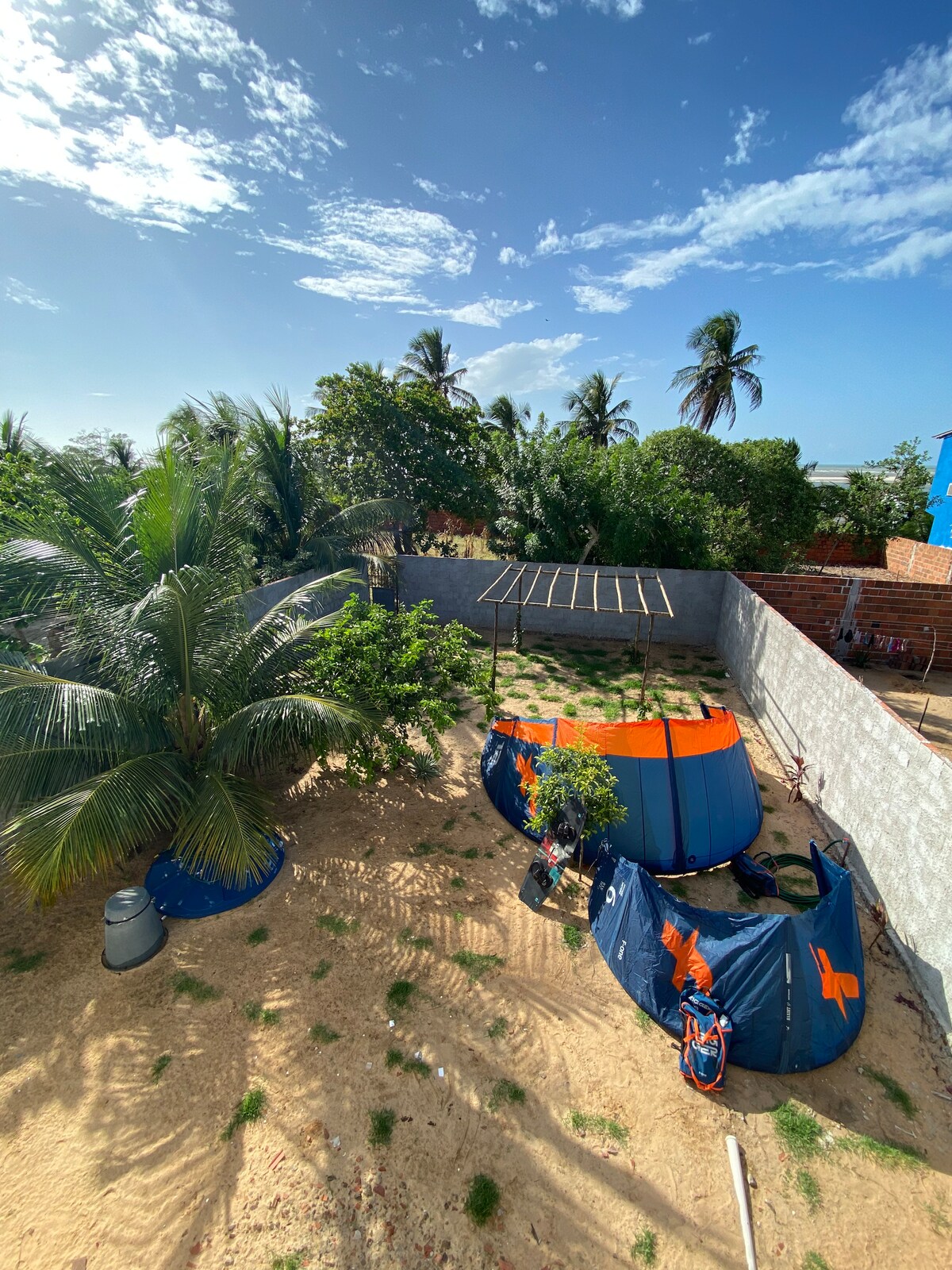 The garden is spacious, featuring sandy ground and various greenery, including palm trees. Two colorful kitesurfing tents are set up, surrounded by small plants and a trellis structure. Bright blue sky and scattered clouds create a light and open atmosphere.