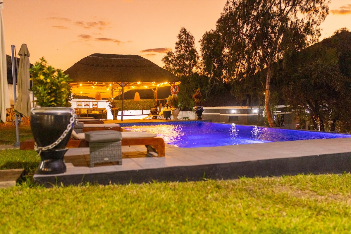 A serene outdoor space features a swimming pool, illuminated with a soft blue light against a warm sunset backdrop. Lounge chairs surround the pool, and a thatched roof structure is visible in the distance, adorned with gentle lighting amidst landscaped greenery.